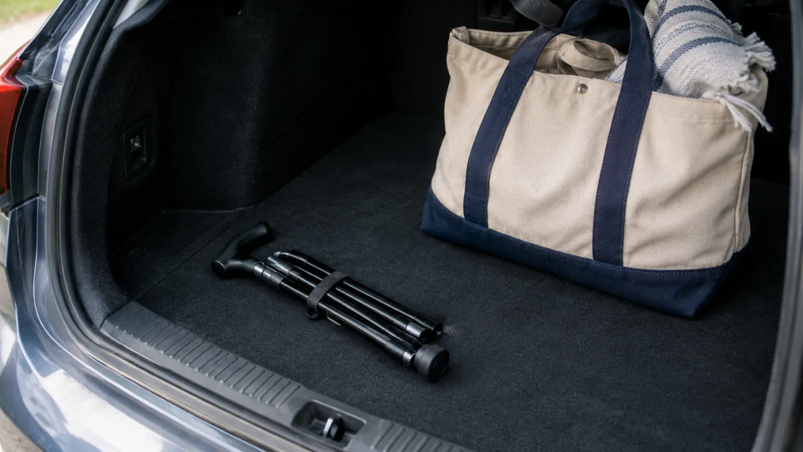 A folding walking cane collapsed into sections resting on the floor of a car trunk beside a tote bag